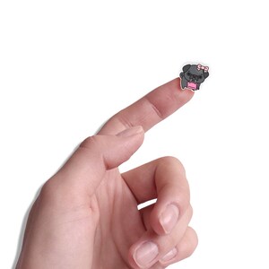 May include: A finger pointing upwards with a small black and white pug sticker with a pink bow and a pink shopping bag on it.