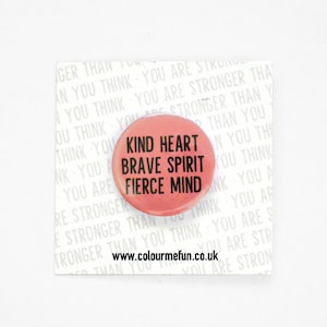 Puede incluir: Insignia de botón rosa con el texto "Kind Heart Brave Spirit Fierce Mind" en letras negras. El sitio web www.colourmefun.co.uk está impreso debajo del texto.