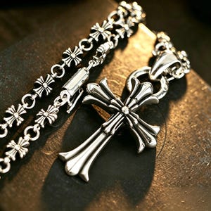 May include: A silver-toned metal necklace featuring a large, ornate cross pendant with flared ends. The chain is composed of repeating small cross-shaped links connected by circular rings, showcasing a polished, reflective metallic finish throughout.