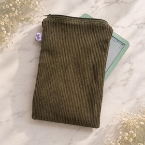 May include: A rectangular olive green corduroy sleeve with a zippered top closure and a small purple fabric tag on the side. A light green electronic tablet is partially inserted into the sleeve, showing a portion of its screen and frame.