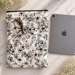 May include: A quilted fabric sleeve featuring a black and white floral pattern with a front pocket. The sleeve has a black zipper closure at the top. A pair of brown-rimmed glasses rests in the front pocket, and a gray tablet is placed beside it.