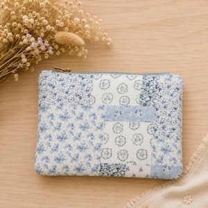 May include: A rectangular fabric pouch featuring a patchwork design of various blue and white floral patterns. The pouch has a gold-toned metal zipper closure along the top edge, providing a secure opening for the interior compartment.