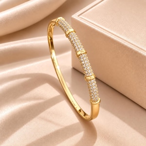 Peut inclure: Un bracelet jonc articulé de couleur dorée avec une section supérieure incurvée incrustée de nombreuses petites pierres claires et scintillantes. Le bracelet présente une finition dorée lisse et polie sur la moitié inférieure.