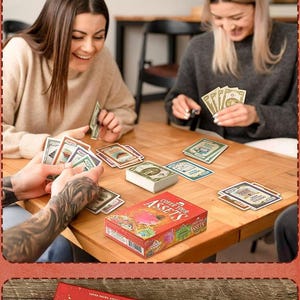 May include: A Cover Your Assets card game set featuring a red box with illustrated graphics and text, a central deck of cards, and multiple individual playing cards with colorful designs and currency values spread across a wooden table surface.