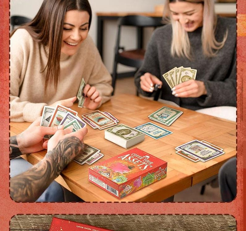 May include: A Cover Your Assets card game set featuring a red box with illustrated graphics and text, a central deck of cards, and multiple individual playing cards with colorful designs and currency values spread across a wooden table surface.