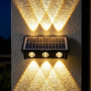 Könnte beinhalten: Eine schwarze, rechteckige Solar-Wandleuchte mit einem oben angebrachten Solarpanel und drei runden LED-Leuchtmitteln. Die Leuchte strahlt warmes Licht in einem Muster nach oben und unten auf die strukturierte Oberfläche der Wand.