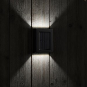 Peut inclure: Une applique murale solaire noire de forme rectangulaire fixée sur une surface en bois verticale. Le luminaire comporte un panneau solaire central et émet deux faisceaux distincts de lumière blanche vive vers le haut et le bas en croix.