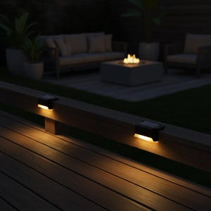 Op de afbeelding: Twee zwarte solar-terraslampen gemonteerd op een houten reling, die een warme, zachte gloed naar beneden werpen op het terrasoppervlak. De lampen hebben een rechthoekig ontwerp met een zonnepaneel aan de bovenkant en een lichtstrip onderaan.