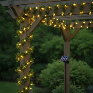 May include: A string of artificial green ivy leaves intertwined with warm white LED fairy lights wrapped around a wooden pergola beam, featuring a small black solar panel mounted on a stake nearby to power the illuminated decorative garland.
