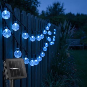 Op de afbeelding: Een lichtsnoer met blauwe led-bollen met een textuur van kleine belletjes, verbonden door een zwarte draad. De lampjes worden gevoed door een vierkant zwart zonnepaneel op een paal, ontworpen om buitenruimtes te verlichten met een blauwe gloed.