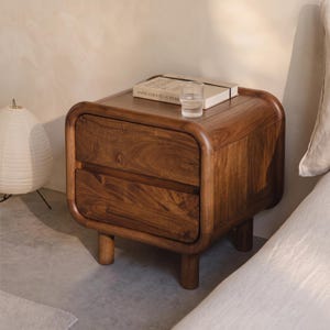 Può includere: Un comodino in legno marrone scuro con angoli arrotondati e due cassetti. Il comodino presenta quattro gambe cilindriche e una finitura liscia. Un libro intitolato Annie Leibovitz at Work e un bicchiere di vetro trasparente sono appoggiati sopra.