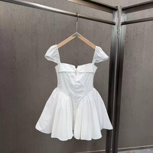 以下が含まれることがあります： スクエアネック、短いフリル袖、中央に小さな白いボタンが縦に並んだフィット感のあるボディスが特徴の白いミニドレス。スカートはボリュームのあるプリーツ仕様で、膝上で広がるフレアなシルエットを作り出しています。