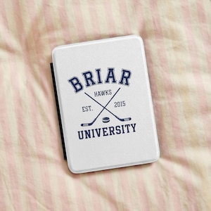 Könnte beinhalten: Eine weiße, rechteckige E-Reader-Hülle mit einer dunkelblauen Grafik und dem Text BRIAR UNIVERSITY HAWKS EST. 2015. Das Design zeigt zwei gekreuzte Eishockeyschläger und einen Puck, die mittig zwischen den Textelementen auf der Vorderseite liegen.