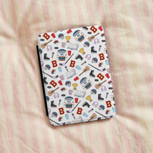 Könnte beinhalten: Eine rechteckige E-Reader-Hülle mit einem wiederkehrenden Muster aus Eishockey-Illustrationen, darunter Trikots mit der Nummer 44, Hockeyschläger, Schlittschuhe, Helme, Pokale, Gitarren, Mikrofone, Kopfhörer, Bücher und der Buchstabe B in Rot.