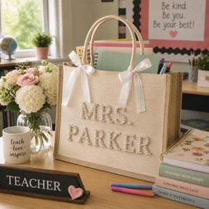 Pode incluir: Uma bolsa tote de juta bege com duas alças adornadas com laços de cetim branco. A frente da bolsa exibe o texto MRS. PARKER formado por contas brancas tipo pérola. A bolsa é retangular com uma base estruturada e topo aberto.