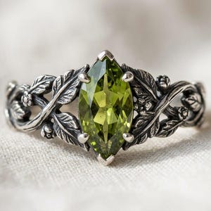 Könnte beinhalten: Ein Ring aus oxidiertem Sterlingsilber mit einem olivgrünen Edelstein im Marquise-Schliff. Die Ringschiene zeigt ein detailliertes Reliefdesign aus ineinander verschlungenen Blättern und kleinen Knospen, die den facettierten Stein einrahmen.