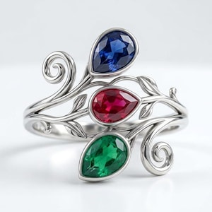 May include: A silver-toned ring featuring a vine-like band with three pear-shaped gemstones in blue, red, and green. The metal band is sculpted into delicate swirling branches and leaves that frame each of the three faceted, colorful stones.