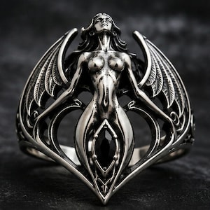 Puede incluir: Un anillo de tono plateado que presenta una figura alada esculpida con cabello largo y mirada hacia arriba. La figura se sitúa sobre una piedra negra central de corte marquesa engastada en un marco de metal calado que forma el anillo.