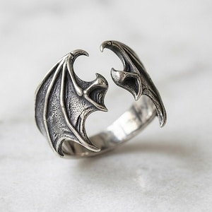 Puede incluir: Un anillo abierto de plata con dos alas detalladas similares a las de un murciélago que se encuentran en el centro. El metal tiene un acabado oscuro y oxidado en las áreas hundidas para resaltar la estructura texturizada de las alas.