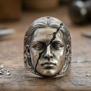 May include: A silver-toned metal ring featuring a sculpted human face with a prominent crack running diagonally across the forehead and nose. The band displays intricate swirling patterns on the sides, finished with a dark, weathered patina.