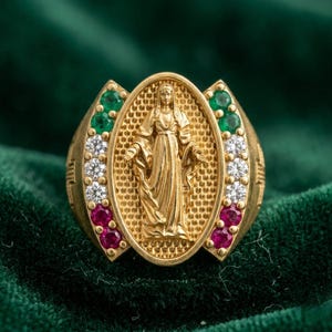 Puede incluir: Un anillo de oro con un medallón ovalado central que presenta un relieve de una figura con túnica. El medallón está flanqueado por dos filas verticales de piedras preciosas, cada una con dos piedras verdes, tres claras y dos rojas a cada lado.