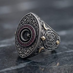 Può includere: Un anello in argento con una parte superiore ovale che presenta un emblema centrale a forma di mezzaluna e stella, circondato da piccole pietre rosse. La fascia è decorata con volute complesse, una stella geometrica e dettagli dorati.