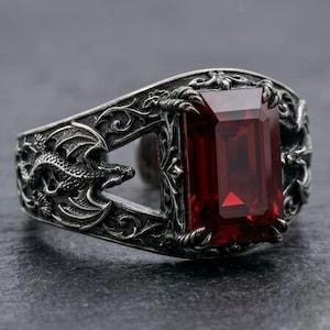 May include: A silver-toned ring featuring an emerald-cut deep red gemstone held by four claw-like prongs. The band is decorated with intricate relief carvings of winged dragons and scrolling filigree patterns across the metal surface.