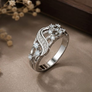 Op de afbeelding: Een zilverkleurige ring met een centrale S-vormige curve ingelegd met kleine heldere steentjes. De ringband is verdeeld in meerdere segmenten, met extra heldere steentjes gezet langs de zijkanten en in het opengewerkte ontwerp van de metalen structuur.