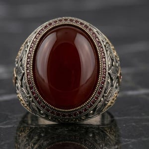 Pode incluir: Um anel de prata com uma grande pedra cabochão oval de cor vermelho-escura no centro. A pedra é rodeada por uma borda fina de pequenas pedras vermelhas. O anel é decorado com padrões de folhas em relevo intrincados e pequenos detalhes dourados.