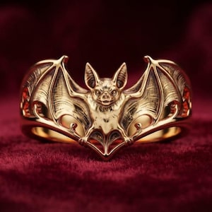 May include: A gold ring featuring a detailed, sculpted bat with outstretched wings. The bat is centered on the band, with its ears pointed upward and wings extending along the sides of the ring, showcasing intricate textures and polished surfaces.
