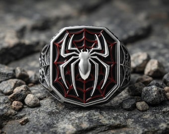 May include: A silver-toned ring featuring an octagonal face with a raised silver spider emblem centered over a black background with red web detailing. The sides of the ring band are engraved with a spiderweb pattern, creating a detailed metallic accessory.