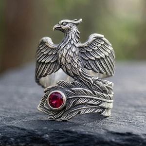 May include: A silver-toned ring featuring a detailed phoenix with spread wings. The bird's tail feathers wrap around the band, incorporating a single, round, deep red gemstone set into the metal. The finish is oxidized to highlight the texture.