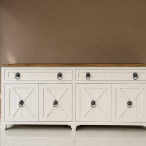 Puede incluir: Un aparador rectangular largo con estructura blanca y superficie superior en tono madera natural. La unidad incluye dos cajones y cuatro puertas de armario, cada una adornada con tiradores oscuros de anilla y detalles de paneles en forma de X.