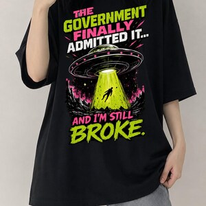 May include: A black t-shirt featuring a graphic print of a flying saucer abducting a figure in a bright green beam of light. The text reads THE GOVERNMENT FINALLY ADMITTED IT... AND I'M STILL BROKE. The design includes pink and green accents.