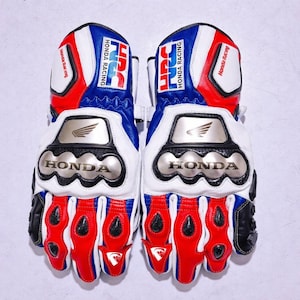 Puede incluir: Un par de guantes de carreras de motos en color rojo, blanco y azul. Los guantes cuentan con protectores metálicos en los nudillos con el logotipo de Honda, la marca HRC y el texto Honda Racing impreso en los puños y dedos con un diseño llamativo.