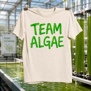 May include: A cream-colored short-sleeve t-shirt featuring the text "TEAM ALGAE" printed in a bright green, textured font. The letters have a distinct organic, leafy appearance, and the shirt is displayed hanging from a wire against a blurred background.