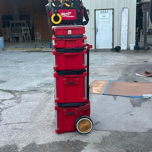 May include: A stack of red and black Milwaukee Packout modular tool storage units. The tower includes a top tool bag, three rigid storage cases, and a rolling base with a handle and two wheels. The Milwaukee logo is visible on the top bag.