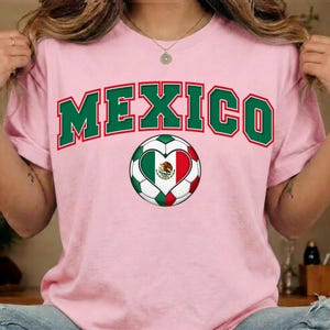 Op de afbeelding: Een lichtroze t-shirt met het woord MEXICO in grote, gebogen, donkergroene blokletters met een rode omlijning. Onder de tekst staat een voetbalafbeelding met een hartvormig midden in de kleuren van de Mexicaanse vlag: groen, wit en rood.