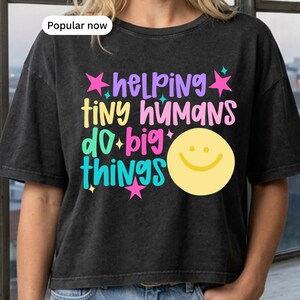 Op de afbeelding: Een antracietgrijs t-shirt met de kleurrijke tekst "helping tiny humans do big things" in paars, geel, roze en turquoise. Het ontwerp bevat kleine stervormen en een groot geel smiley-icoon aan de rechterkant van de afbeelding.