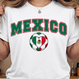 Op de afbeelding: Een wit t-shirt met het woord MEXICO in grote, gebogen groene letters met een rode omlijning. Onder de tekst staat een voetbalafbeelding met een hartvormig middenstuk dat de groen-wit-rode Mexicaanse vlag met het centrale embleem bevat.