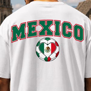 Op de afbeelding: Een wit t-shirt met het woord MEXICO in grote groene letters met een rode omlijning in een boogvorm. Onder de tekst staat een voetbalafbeelding met een hartvormig midden in de kleuren van de Mexicaanse vlag: groen, wit en rood.