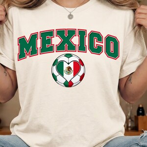 Op de afbeelding: Een cr&egrave;mekleurig T-shirt met het woord MEXICO in een groot, gebogen groen lettertype met een rode omlijning. Onder de tekst staat een voetbalafbeelding met een hartvormig midden dat de groen-wit-rode Mexicaanse vlag toont.