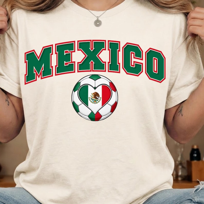 Op de afbeelding: Een cr&egrave;mekleurig T-shirt met het woord MEXICO in een groot, gebogen groen lettertype met een rode omlijning. Onder de tekst staat een voetbalafbeelding met een hartvormig midden dat de groen-wit-rode Mexicaanse vlag toont.