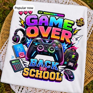 May include: A white t-shirt featuring a colorful graphic with the text GAME OVER BACK TO SCHOOL. The design includes a black game controller, headphones, a calculator, a backpack, a computer mouse, a notebook, and a level up icon.