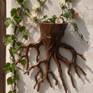Op de afbeelding: Een bruine wandplantenbak in de vorm van een boomstronk met blootliggende, uitlopende wortels. De plantenbak heeft een gladde, glanzende afwerking en bevat een kleine groene plant met lichtroze en witte bloemen tegen een witte muur.