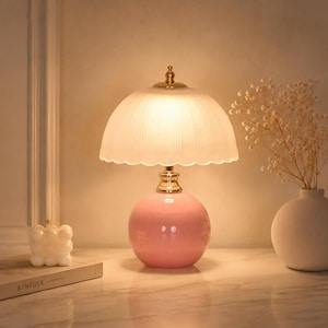 May include: A table lamp featuring a glossy pink spherical ceramic base and a scalloped, frosted white glass shade. The lamp has a gold-toned metal neck and emits a warm, soft glow, illuminating the surrounding area with a gentle light.