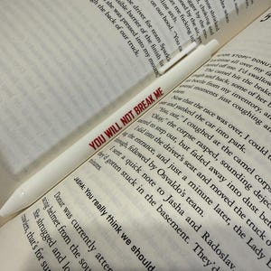 May include: A white pen with the text YOU WILL NOT BREAK ME printed in red ink, resting diagonally across an open book with black printed text on white pages. The pen has a smooth, cylindrical body and a pointed tip.
