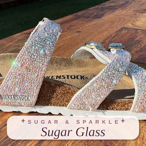 May include: A Birkenstock sandal featuring two straps covered in iridescent, light-reflecting crystals. The footbed is made of cork with a white sole. The text Sugar & Sparkle Sugar Glass is visible on a light-colored rectangular overlay at the bottom.