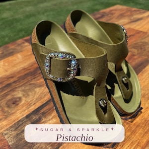 May include: A pair of olive green thong sandals featuring a cork footbed and a large, rectangular buckle encrusted with iridescent crystals. The sandals have a toe post design and the text SUGAR & SPARKLE Pistachio is printed on a label.
