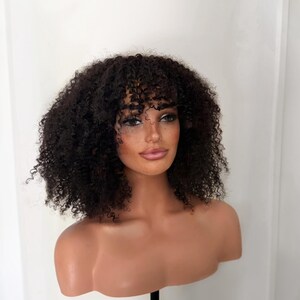 May include: A dark brown wig with tight, voluminous curls and a fringe that rests above the eyebrows. The hair is styled in a shoulder-length cut, displayed on a smooth, light-toned mannequin head and shoulders against a plain white background.